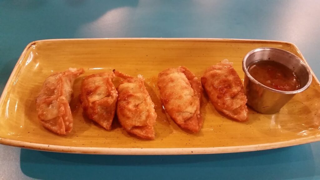Five golden-brown fried dumplings are neatly arranged in a row on a rectangular yellow plate. A small metal pot filled with dipping sauce sits at the right end of the plate. The background is a smooth blue tabletop.