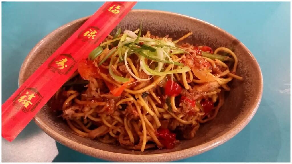 A brown bowl filled with stir-fried noodles, shredded meat, red pepper, and spring onion strips is on a blue table. Bright red chopsticks with gold Chinese characters rest on the bowl's rim. The dish looks colourful and appetising.