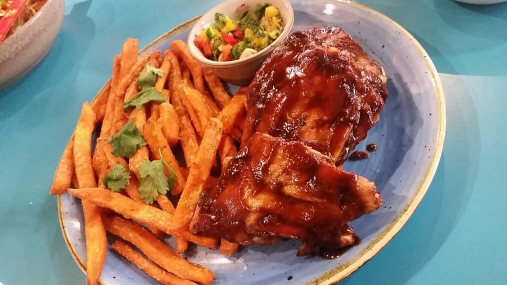 A blue plate with glazed barbeque ribs, sweet potato chips garnished with coriander, and a small pot of chunky salsa made of diced tomatoes, onion, and herbs. The food is served on a blue table.