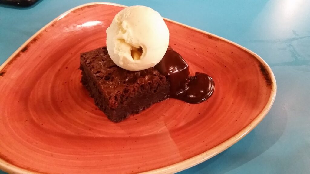 A scoop of vanilla ice cream sits atop a square chocolate brownie with chocolate sauce on the side, served on a triangular, orange ceramic plate placed on a blue table.