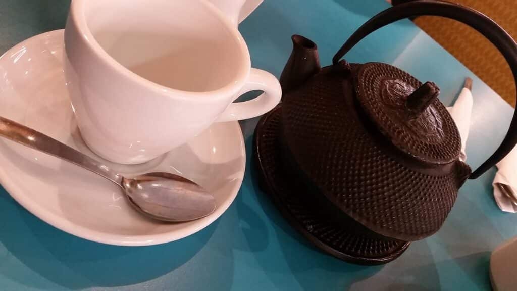 A white teacup on a saucer with a metal spoon sits next to a black, textured cast iron teapot on a blue table. A folded white serviette and part of a chair are visible in the background.