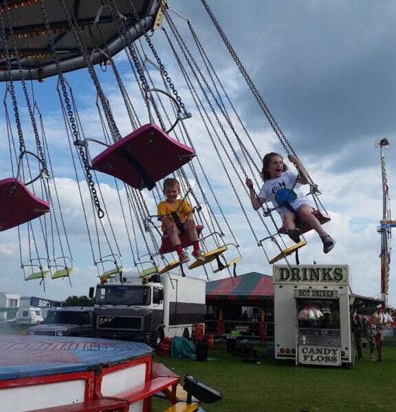 Two children ride colourful swings at a fairground. The girl on the right smiles and grips the chains, whilst the younger child on the left looks forward. Fair stalls, lorries, and a cloudy sky are visible in the background. The ground is grassy.