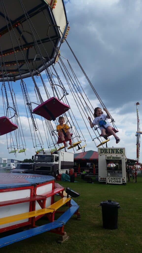 Two children sit on colourful swing seats suspended by chains, smiling as they ride a spinning funfair swing ride outdoors. The sky is partly cloudy, and funfair stalls, lorries, and a "DRINKS" stand are visible in the background on grassy ground.