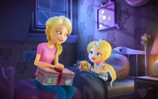 In a cosy, purple-themed bedroom, a woman with blonde hair in a plait smiles at a little blonde girl in pyjamas sitting on a bed with a puppy. The woman holds a decorated box. The room features heart-shaped wall patterns and a window showing a night sky.