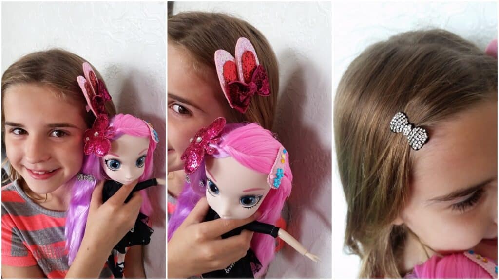 A girl with light brown hair and a grey-striped shirt smiles whilst holding a doll with pink and purple hair. Both wear shiny butterfly hair clips. A close-up shows her hair with a sparkly rhinestone bow clip against a white wall.