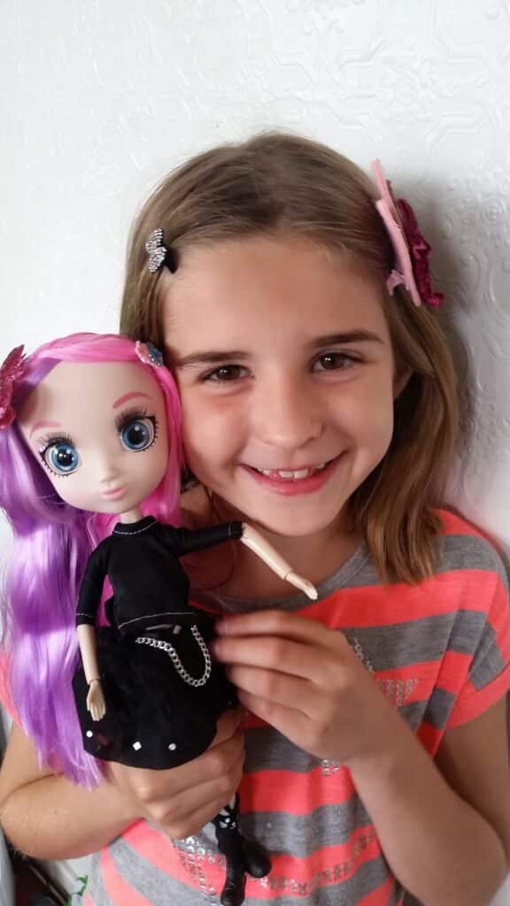 A smiling young girl with straight brown hair, wearing a grey and pink striped shirt, holds a doll with long purple hair, wide blue eyes, and a black dress. Both have hair clips, and they stand against a white textured wall.