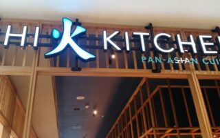 A restaurant sign reads “KITCHEN” with a stylised blue symbol and partial letters “CHI” on the left. Below, smaller blue letters say "PAN-ASIAN CUISINE." The background features wooden frames and ceiling lights inside the building.