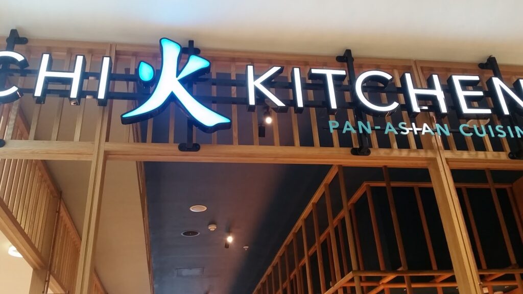 A restaurant sign reads "CHI KITCHEN" in bold, modern letters with a blue stylised logo between "CHI" and "KITCHEN." Below, smaller text says "PAN-ASIAN CUISINE." The background features wooden slats and an open ceiling design.