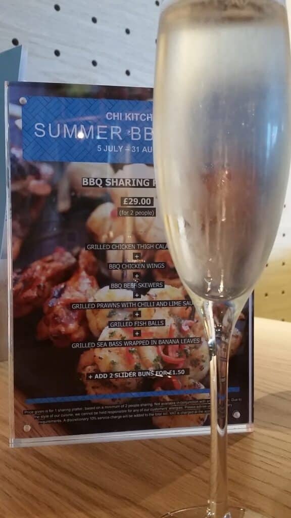 A glass of sparkling wine is in focus in the foreground. Behind it is a menu advertising a “Summer BBQ” at Chi Kitchen, listing BBQ sharing options and prices, with images of grilled meats as a colourful background. The scene is set indoors.