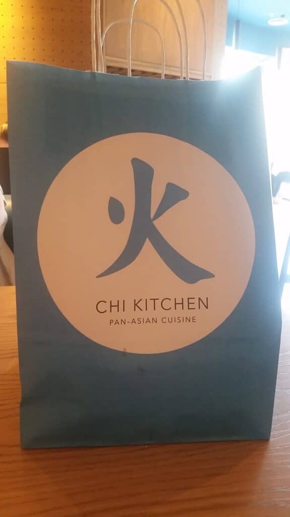 A blue paper bag with a white circle featuring a large Asian character, below which reads “CHI KITCHEN” and “PAN-ASIAN CUISINE,” sits on a wooden table in a softly lit indoor setting. Handles are visible at the top of the bag.