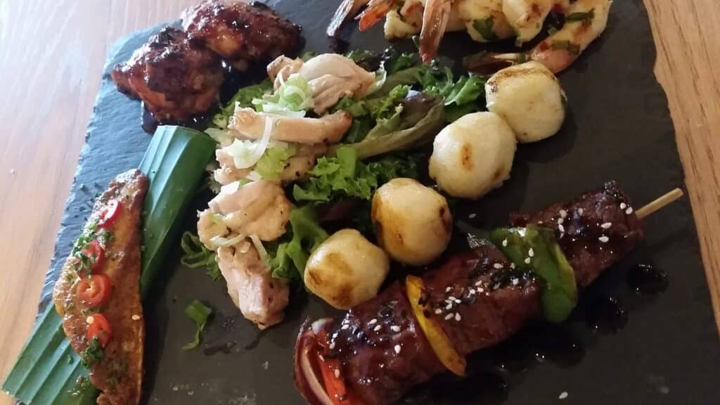 A slate platter features grilled chicken, prawn skewers, grilled beef skewer with sesame seeds, crispy round potatoes, sliced chicken on salad leaves, and a spiced grilled sausage garnished with chopped herbs and chilli, arranged artfully on a wooden table.
