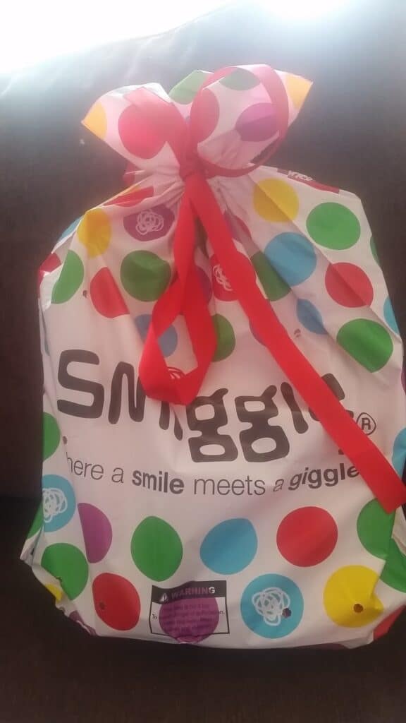 A colourful gift bag with multicoloured polka dots, red ribbon tied at the top, and the text “Snuggie—where a smile meets a giggle” printed on the front, sits on a dark sofa. A purple warning label is visible near the bottom.