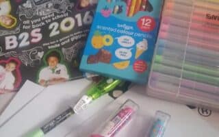 A variety of school supplies including three colourful notebooks, a pack of scented coloured pencils, a set of multicoloured pens, glue sticks, highlighters, and several white envelopes, all laid out on a patterned surface.