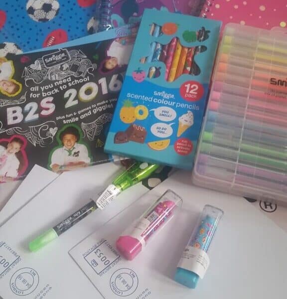 A variety of school supplies including three colourful notebooks, a pack of scented coloured pencils, a set of multicoloured pens, glue sticks, highlighters, and several white envelopes, all laid out on a patterned surface.
