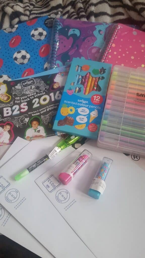 A collection of colourful school supplies including notebooks with various patterns, a pack of scented coloured pencils, pastel highlighters, gel pens, glue sticks, and several envelopes or papers labelled “B2S 2019” spread out on a surface.
