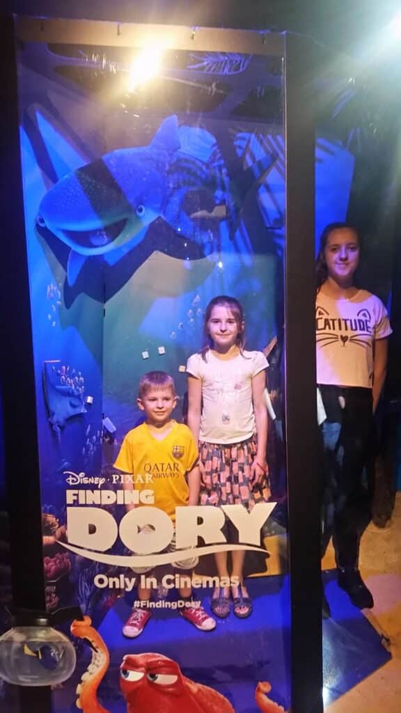 Three children stand inside a display for the film "Finding Dory." Two are inside the frame with underwater ocean graphics and Dory characters behind them, while one stands just outside. The film title and release info are at the bottom of the transparent panel.