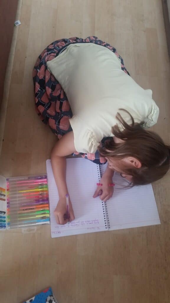 A young girl with light brown hair, wearing a cream top and a patterned skirt, sits on a wooden floor, leaning over a spiral notebook. She writes in purple pen. Beside her is a clear case containing colourful pens arranged in a row.