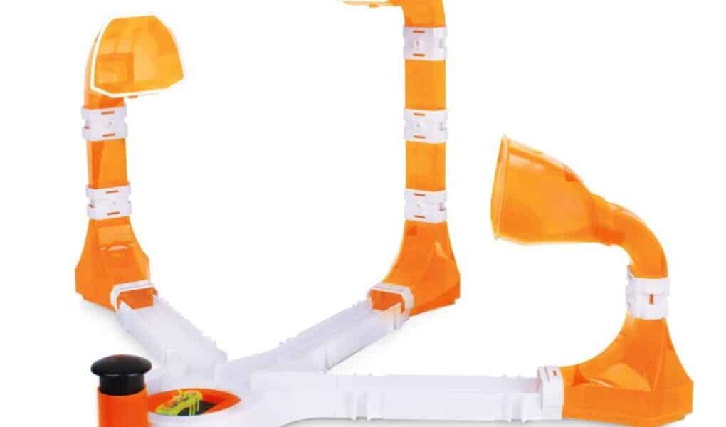 A plastic game with three orange and white ramps, each ending in a dome-shaped cup, radiating from a central base. One side has a black plunger for launching a small ball towards the cups. The design is open, colourful, and for tabletop play.