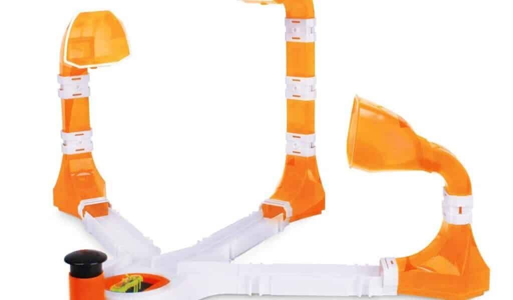 A plastic game with three orange and white ramps, each ending in a dome-shaped cup, radiating from a central base. One side has a black plunger for launching a small ball towards the cups. The design is open, colourful, and for tabletop play.