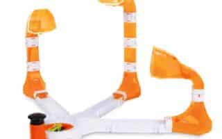 A plastic game with three orange and white ramps, each ending in a dome-shaped cup, radiating from a central base. One side has a black plunger for launching a small ball towards the cups. The design is open, colourful, and for tabletop play.