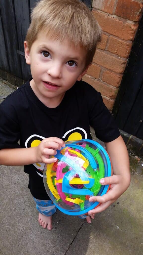 A young boy with light brown hair stands barefoot on concrete near a brick wall, looking up. He wears a black shirt with a yellow and white design and holds a colourful, spherical maze puzzle toy in his hands.
