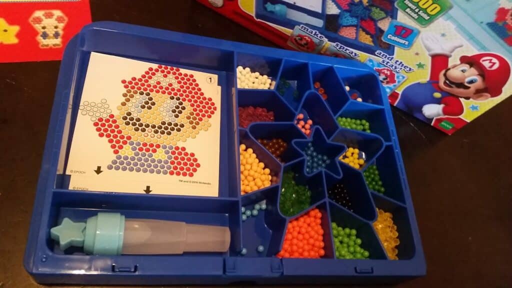 A craft kit tray holds coloured beads sorted into compartments shaped like a star. A Mario design guide with a beaded pixel art image is placed in the tray. A blue tool with a star-shaped top is included. Mario is pictured on the product box in the background.