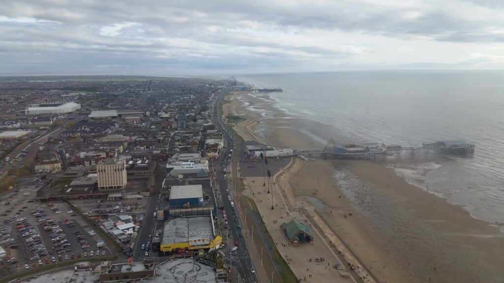 Aerial view of a coastal town with a sandy beach, a pier extending into the sea, and a main road running parallel to the shoreline. Buildings, car parks, and scattered people are visible under a mostly cloudy sky.