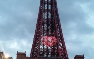The Blackpool Tower stands tall under a cloudy sky. Its iron structure is illuminated, with a glowing heart-shaped light in the centre. The lower part shows the red-bricked base and the text “THE BLACKPOOL TOWER” above the entrance.