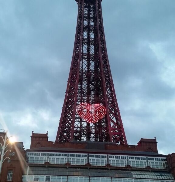The Blackpool Tower stands tall under a cloudy sky. Its iron structure is illuminated, with a glowing heart-shaped light in the centre. The lower part shows the red-bricked base and the text “THE BLACKPOOL TOWER” above the entrance.