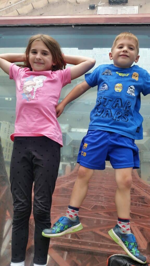 Two children pose in front of a glass window. The girl on the left wears a pink shirt and black trousers with polka dots, and has her hands behind her head. The boy on the right wears a blue Star Wars shirt, blue shorts, and colourful trainers.