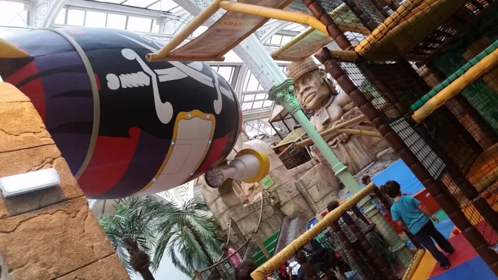 Indoor play area with a pirate theme: a large pirate ship floats above, decorated with a skull and crossbones. There are rope climbing structures, a pirate statue, artificial palm trees, and children playing on colourful mats under a glass ceiling.