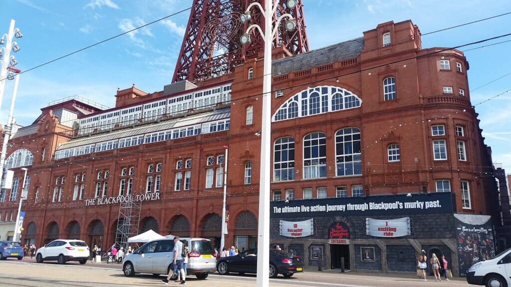 Red-brick façade of Blackpool Tower under a blue sky, with people and parked cars in front. A banner advertises “The ultimate live action journey through Blackpool’s murky past.” Tram wires cross above the street.