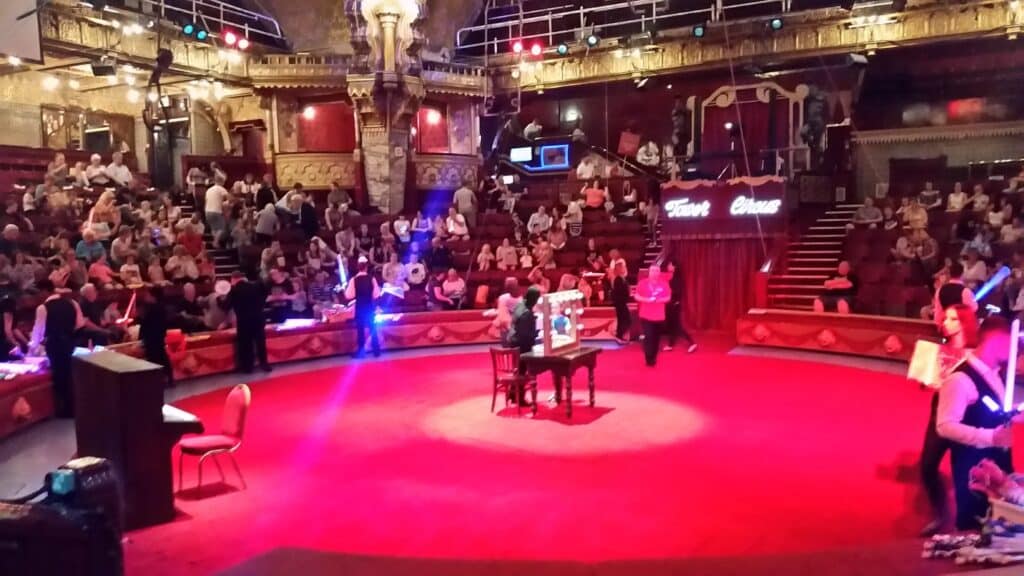 A brightly lit circus ring with a table and chairs in the centre, surrounded by an audience in tiered seats. Performers with glowing props interact on the red floor, while ornate decorations and a sign reading "Tower Circus" are visible in the background.