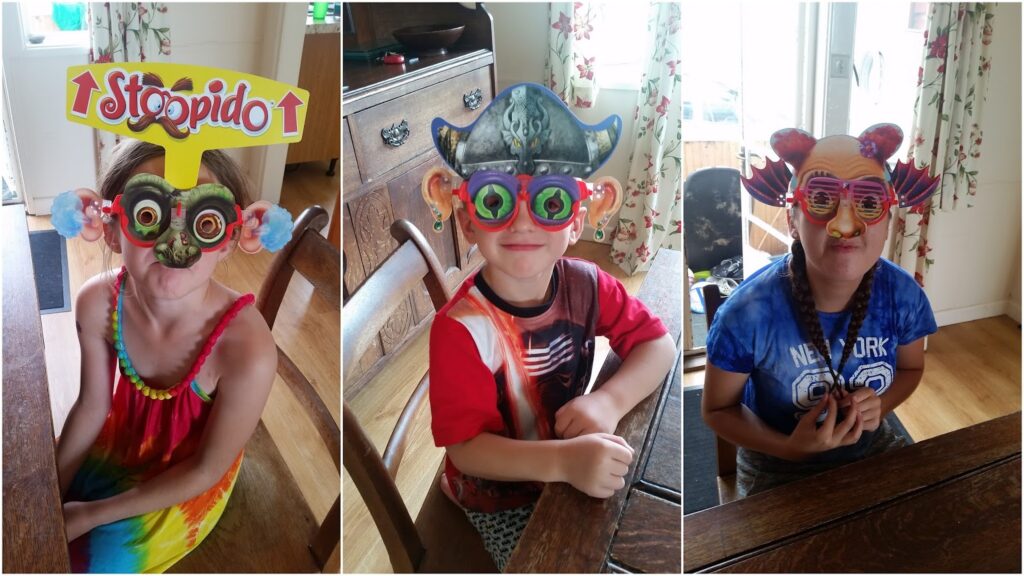 Three children sit indoors, each wearing colourful, silly masks and glasses. The first child’s mask says “Stoopido,” the second wears a pirate hat and funny glasses, and the third has a cat-themed mask. All are smiling and seated at a wooden table.