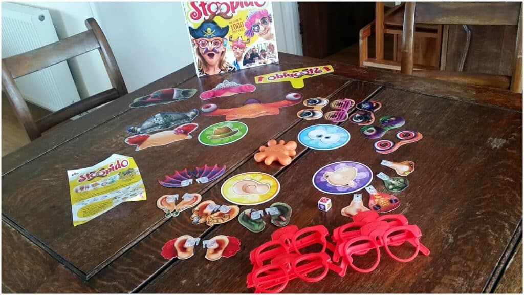 A wooden table with the board game "Stoopido" set up. Various colourful cardboard face pieces, a die, plastic red glasses, playing cards, and game instructions are spread out, with the game box standing upright in the background.