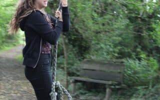 A young woman with long brown hair, dressed in a black jacket, trousers, and trainers, holds onto a thick rope swing outdoors. She stands on a gravel path surrounded by lush green trees. A wooden bench sits in the background among the foliage.