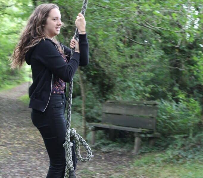 A young woman with long brown hair, dressed in a black jacket, trousers, and trainers, holds onto a thick rope swing outdoors. She stands on a gravel path surrounded by lush green trees. A wooden bench sits in the background among the foliage.