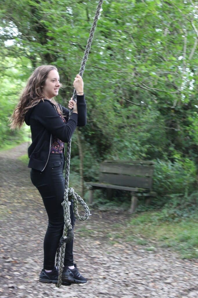 A young woman with long brown hair, wearing a black jacket, black trousers, and black trainers, holds onto a thick rope swing in a wooded outdoor area. There is a wooden bench and dense green foliage in the background.