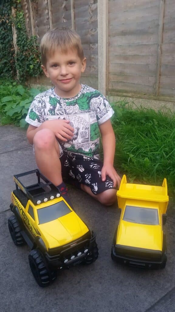 A young boy with fair hair sits outdoors on the pavement, smiling. He wears a white and green shirt with black patterned shorts. In front of him are two large yellow toy lorries. Grass and a wooden fence are visible in the background.