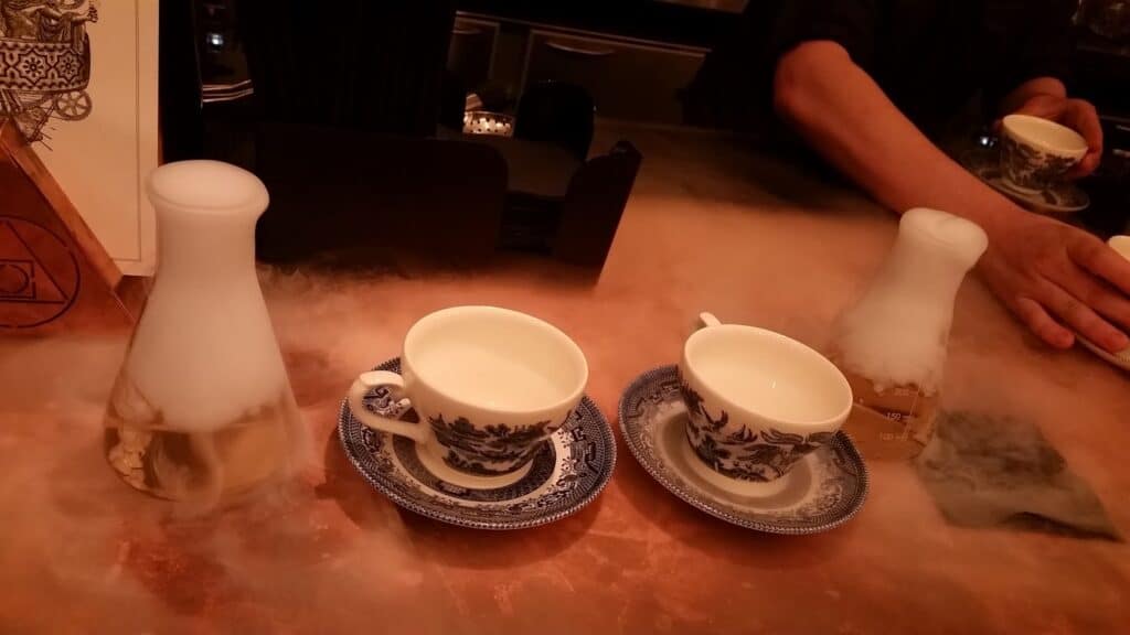 Two ornate teacups and saucers sit on a copper worktop beside two glass flasks with white vapour spilling out. A bartender in black serves another cup in the background. The scene suggests a creative or molecular cocktail presentation.