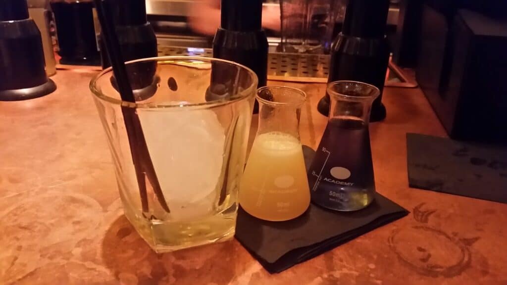 A glass with a large ice sphere and two black straws sits next to two small lab beakers on a serviette, one filled with yellow liquid and the other with purple liquid, on a copper-toned bar counter with bar equipment in the background.