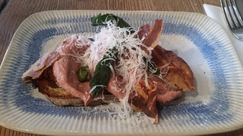 A slice of toasted bread topped with pink pâté, crispy bacon rashers, grilled courgette, and a generous amount of finely grated cheese, served on a rectangular plate with a blue border. A fork and serviette are placed to the side.