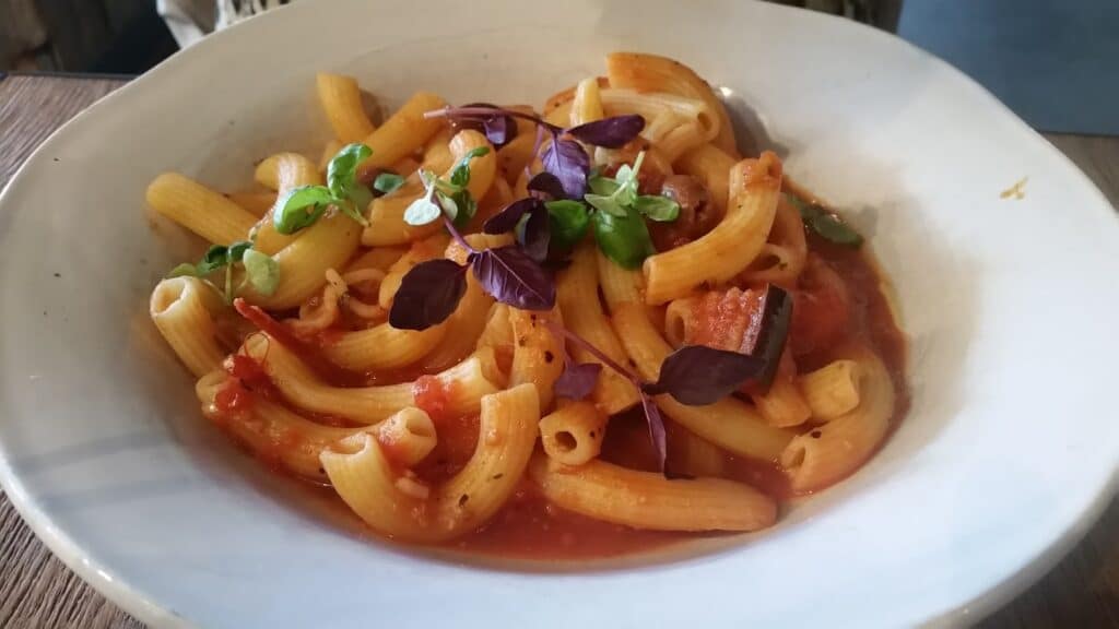 A white bowl filled with rigatoni pasta in a light tomato sauce, garnished with small green and purple microgreens, sits on a wooden table. The dish appears simple and fresh, with no cheese or heavy toppings visible.