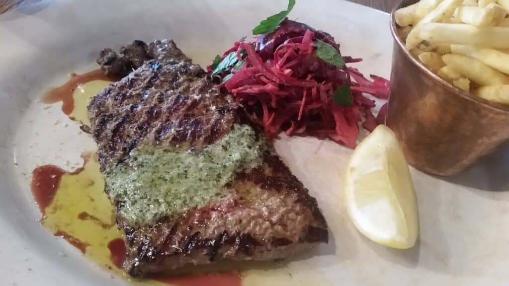 A grilled steak with a green herb butter topping is served with a portion of golden chips in a copper cup, a small mound of shredded beetroot salad garnished with mint, and a lemon wedge on a white plate.