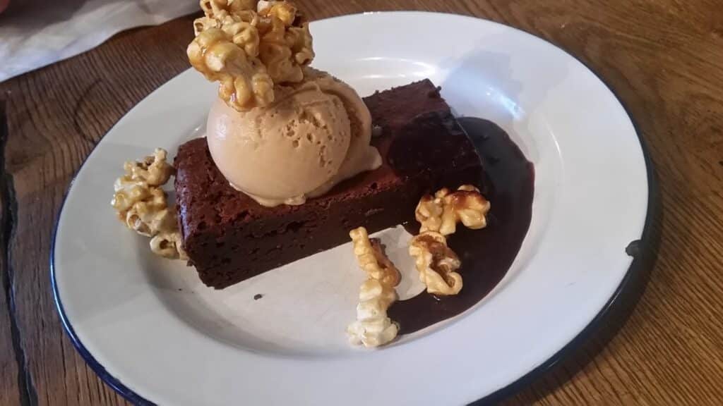 A rectangular slice of chocolate brownie topped with a scoop of ice cream and caramel popcorn, served on a white plate with a dark drizzle of chocolate sauce, placed on a wooden table.