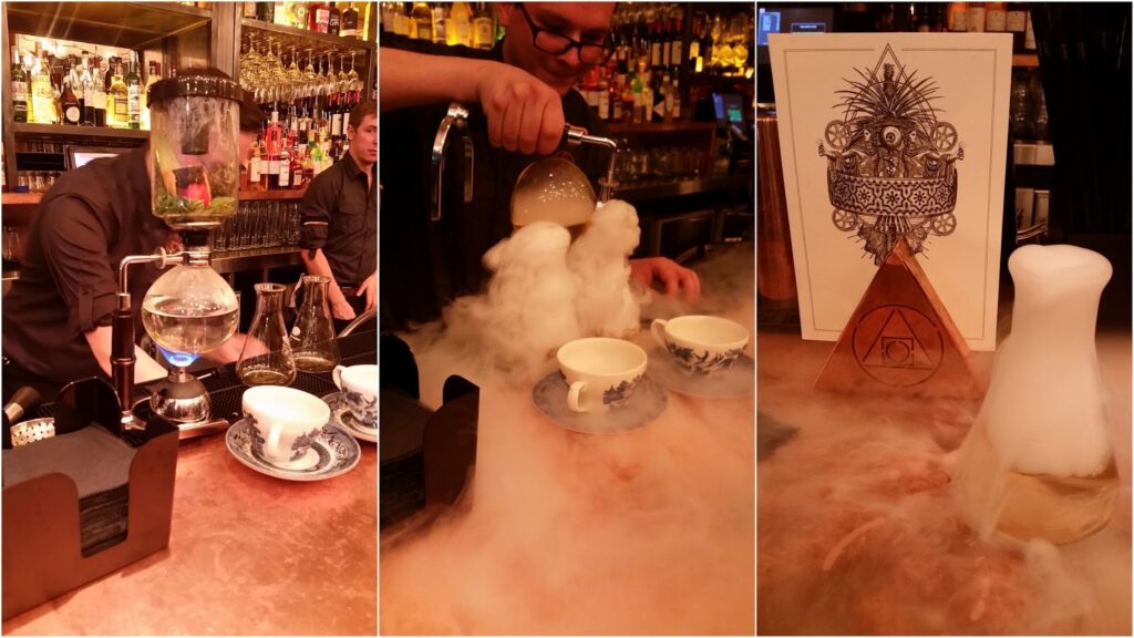 A barman prepares a smoking cocktail using a siphon and glassware, then pours the drink into teacups, creating thick clouds of fog that spread across the bar. A decorative menu with intricate art and mystical symbols stands nearby.