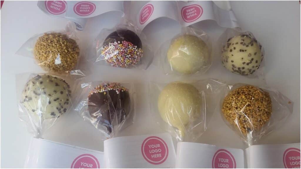 Eight chocolate cake pops, each wrapped in clear plastic, are arranged in two rows. They are variously coated: some with chopped nuts, others with chocolate chips or rainbow sprinkles. Each has a white stick with a label reading, “YOUR LOGO HERE” in pink.