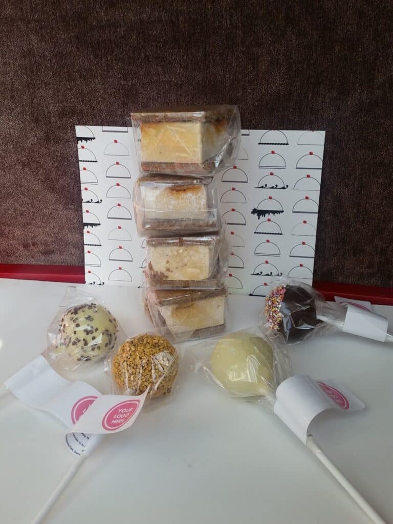 Four cake pops, each coated with different toppings such as sprinkles and nuts, are displayed in front of five stacked packaged cake slices. The background features a card decorated with minimalist cupcake illustrations.