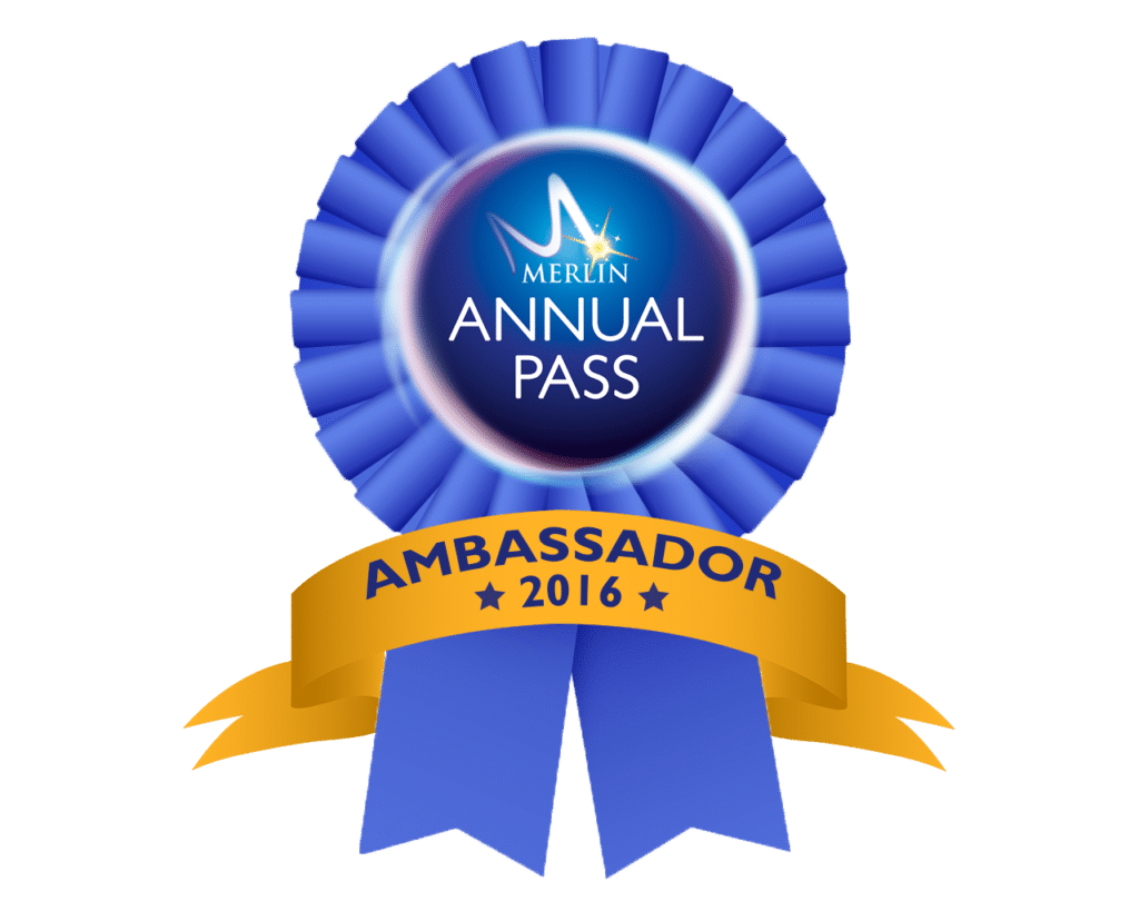 A blue rosette ribbon features a central circle with "MERLIN ANNUAL PASS" in white over a dark blue background. A gold banner below reads "AMBASSADOR 2016" with black stars, and two blue ribbon tails hang down.