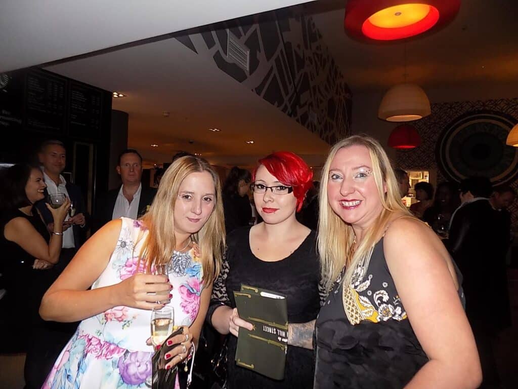 Three women pose at a social event, dressed in semi-formal attire. The woman on the left wears a floral dress and holds a drink. The woman in the centre has bright red hair and glasses, holding a notebook. The woman on the right wears a black dress. Other people mingle in the background.